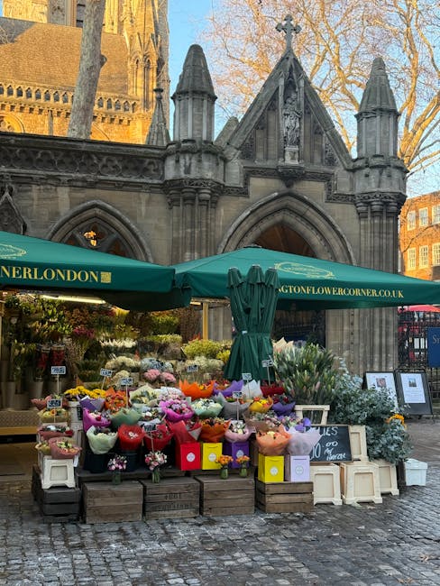 A vibrant outdoor flower stall set against a historic stone archway in a city square, featuring a wide array of fresh flowers arranged in bouquets wrapped in pastel-colored paper and tissue, including